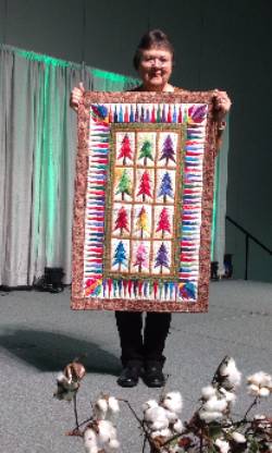 Woman standing on a stage with cotton plant in front of her that has  white cotton balls on it. Woman is holding a quilted wall hanging that has twelve blocks with a Christmas tree in various colors in the center of each block. Cone shapes in various colors border the blocks. Dark printed fabric is the outside border.